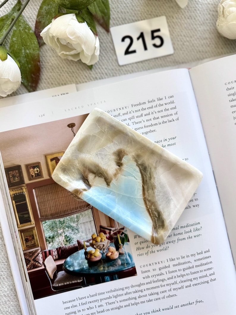 May include: A rectangular, polished stone with a light blue and brown marbled pattern. The stone is resting on an open book with the number "215" visible in the background. The book's pages display text about meditation and self-care.