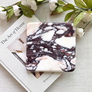 Peut inclure: Un dessous de verre carré en marbre avec un motif blanc et bordeaux, posé sur un livre intitulé "The Art of Home". Des fleurs blanches et des feuilles vertes sont disposées autour du dessous de verre et du livre.