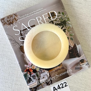 May include: A pale yellow, circular decorative bowl with a hollow center. The bowl is resting on a book titled "SACRED" by Carley Summers. The book is open, displaying images of home decor. A white label with the text "A422" is in the bottom right corner.