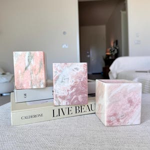 May include: Three square, marble-like decorative containers in shades of pink and white. The containers are arranged on top of books with the text "LIVE BEAUTY" visible. The overall aesthetic is minimalist and elegant.