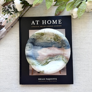 May include: A round coaster with a watercolour design in blue, green, and pink sits on a black book titled "AT HOME". The book cover features the author's name, Brian Paquette. White flowers and green leaves are in the background.