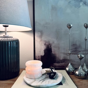 May include: A round white marble tray with grey veining holds a stack of three pale pink translucent glass rings and a set of car keys. The tray rests on a book titled Live Beautiful by Calderone, positioned next to a dark green ribbed lamp.