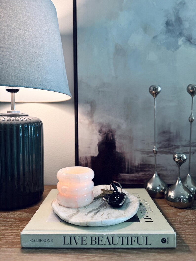 May include: A round white marble tray with grey veining holds a stack of three pale pink translucent glass rings and a set of car keys. The tray rests on a book titled Live Beautiful by Calderone, positioned next to a dark green ribbed lamp.