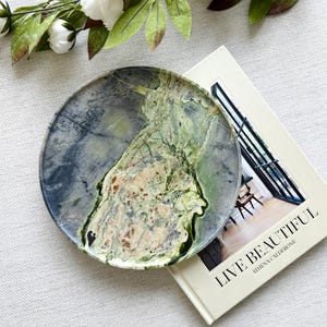 Op de afbeelding: Een ronde decoratieve plaat met een gemarmerd ontwerp in groen, grijs en beige tinten. De plaat ligt op een boek met de titel "LIVE BEAUTIFUL" van Athena Calderone, met een bloemstuk op de achtergrond.