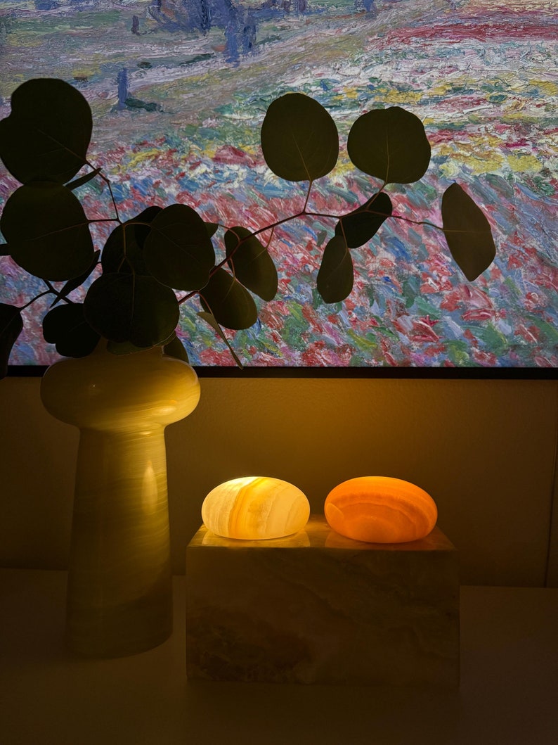 May include: A decorative arrangement featuring two glowing, oval-shaped stone lamps in shades of yellow and orange, resting on a rectangular stone base. A tall, light-colored vase with a sprig of eucalyptus is to the left.