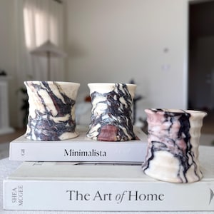 May include: Three decorative marble vases with a unique cylindrical shape. The vases feature a cream base with striking patterns of dark brown and burgundy veining. The vases are displayed on top of two books with the titles "Minimalista" and "The Art of Home".
