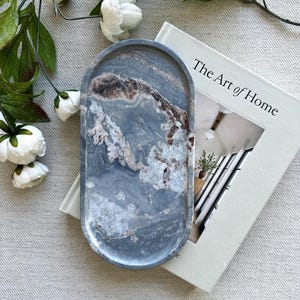 May include: An oval-shaped tray with a smooth, polished surface featuring a marbled pattern in shades of slate gray, white, and brown. The tray is positioned on top of a book titled The Art of Home, accompanied by white floral stems.
