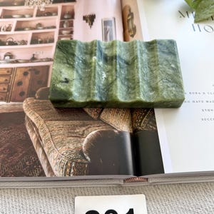 May include: A rectangular, green soap dish with a wavy design. The dish is made of a stone-like material and is resting on an open book. The number "201" is printed on a white tag.