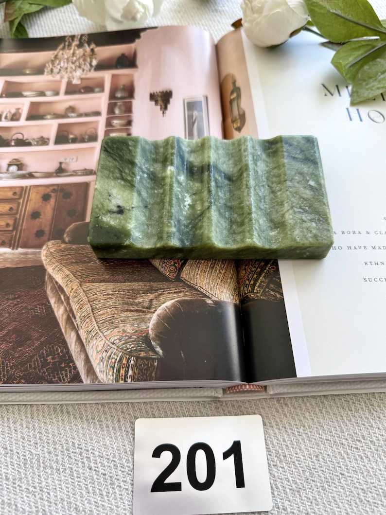 May include: A rectangular, green soap dish with a wavy design. The dish is made of a stone-like material and is resting on an open book. The number "201" is printed on a white tag.