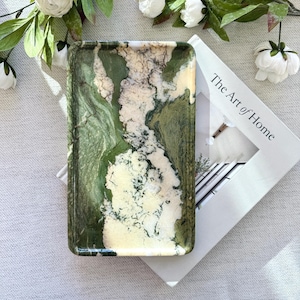 May include: A rectangular decorative tray with a marbled pattern in shades of green and white. The tray is resting on a book with the text "The Art of Home". White flowers and green leaves are arranged around the tray.