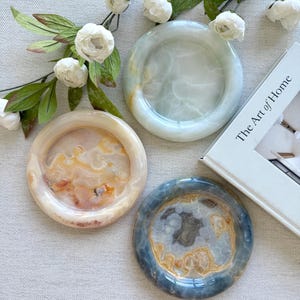 May include: Three circular stone trays with polished surfaces. One tray is translucent white, one is cream with orange swirls, and one is dark blue with grey and gold patterns. A white book titled The Art of Home and a white floral branch are present.