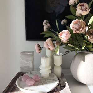 May include: A white marble tray holding three smooth pink rose quartz stones, placed on a book titled Pacific Natural At Home by Jenni Kayne. Nearby are two white ribbed pillar candles, a beige textured vase with pink flowers, and a marble block.