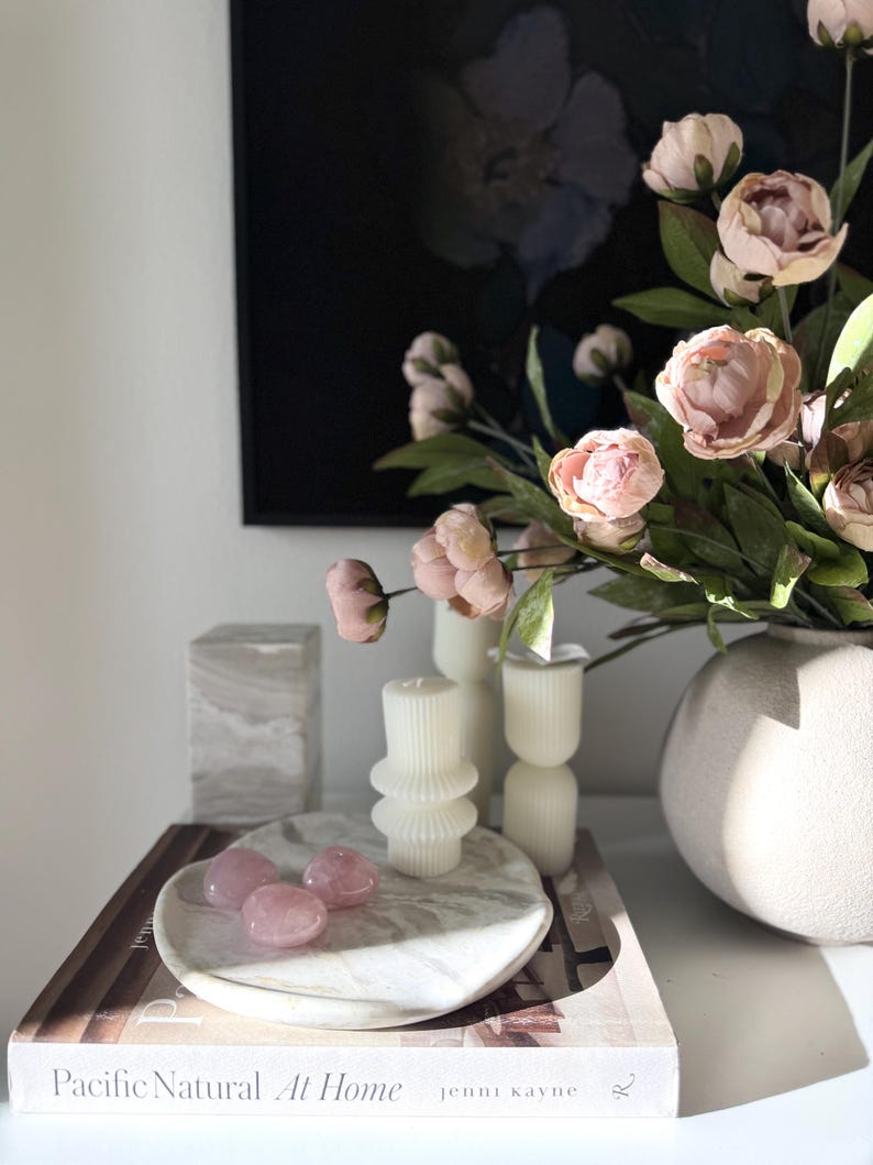 May include: A white marble tray holding three smooth pink rose quartz stones, placed on a book titled Pacific Natural At Home by Jenni Kayne. Nearby are two white ribbed pillar candles, a beige textured vase with pink flowers, and a marble block.
