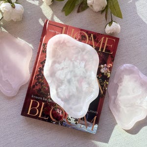 Peut inclure: Trois plateaux translucides rose pâle de forme organique avec une finition lisse et brillante. Chaque plateau présente un bord irrégulier et ondulé ainsi qu'un centre légèrement concave, rappelant l'aspect de la pierre polie ou de la résine.