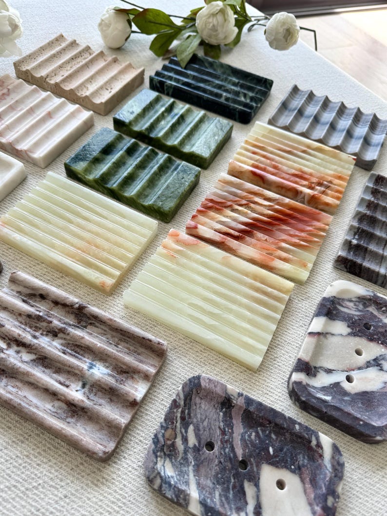 May include: A collection of various soap dishes made of stone. The dishes are rectangular with a grooved design to allow for drainage. Colors include beige, green, white, and marble patterns. Some have a wavy design.