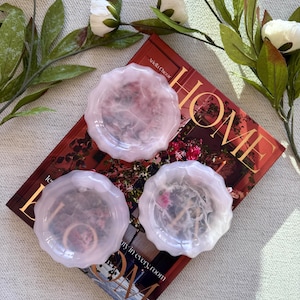 May include: Three round, scalloped-edge resin coasters in a translucent pale pink hue. Each coaster contains embedded dried floral elements and small gold-toned letter charms. They are arranged on top of a red hardcover book titled HOME.