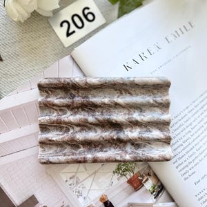 May include: A rectangular soap dish with a wavy design. The dish is a mix of brown and white, resembling marble. The number "206" is visible in the background, along with a magazine.
