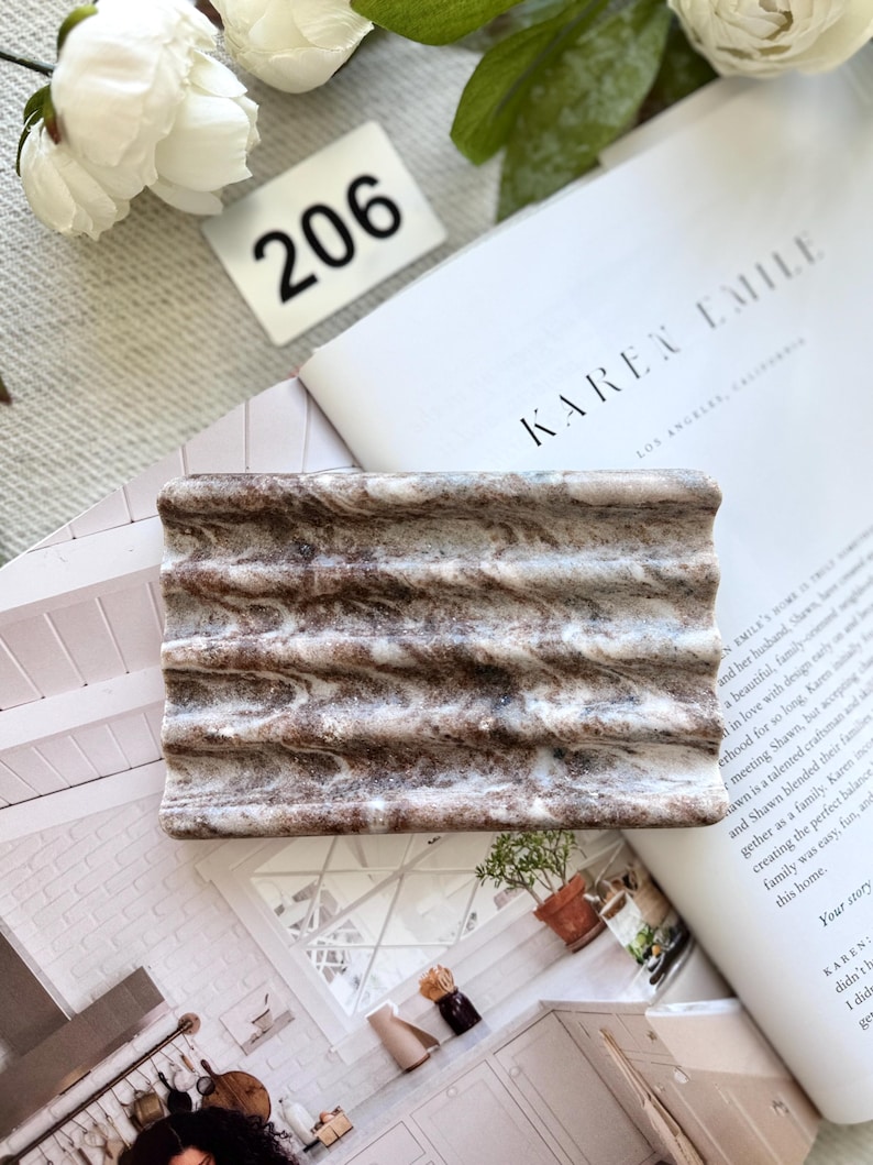 May include: A rectangular soap dish with a wavy design. The dish is a mix of brown and white, resembling marble. The number "206" is visible in the background, along with a magazine.