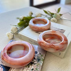 May include: Three round, translucent bangles with a marbled effect in pink and red hues. The bangles are displayed on a book and magazine, with white flowers. The bangles have a smooth, polished finish. Each bangle is approximately 7 cm in diameter.