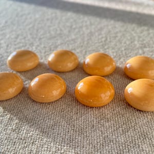 May include: Eight smooth, oval-shaped, amber-colored stones are arranged on a neutral-toned, textured surface. The stones have a glossy finish, reflecting light. The arrangement suggests a collection of decorative or natural elements.