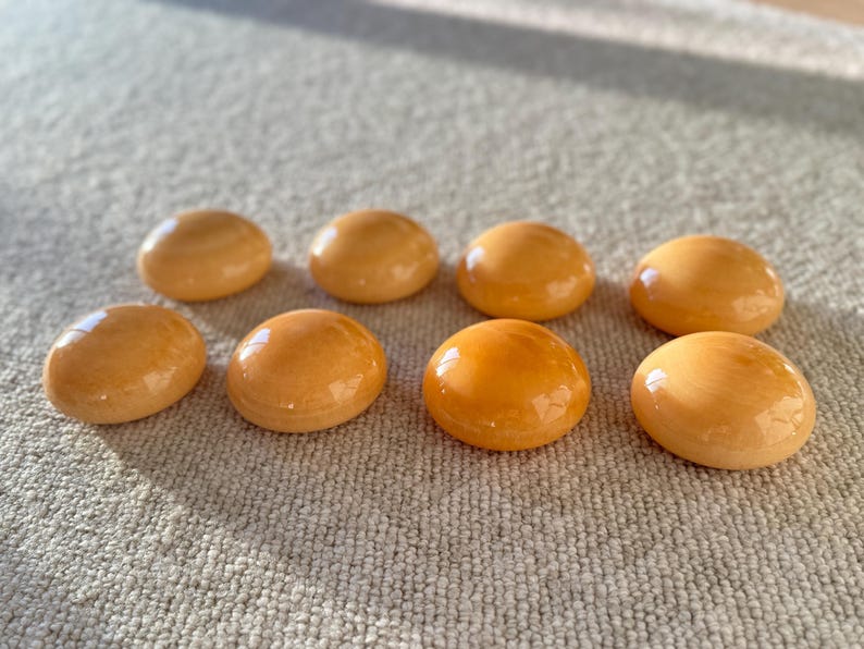 May include: Eight smooth, oval-shaped, amber-colored stones are arranged on a neutral-toned, textured surface. The stones have a glossy finish, reflecting light. The arrangement suggests a collection of decorative or natural elements.