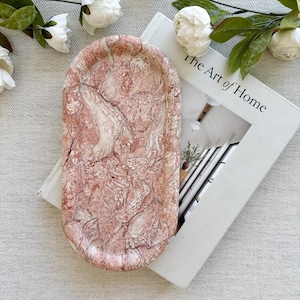 May include: An oval-shaped tray featuring a textured, marbled surface with swirling patterns of dusty rose, cream, and white. The tray has a raised rim and a smooth, polished finish, displaying a complex, organic stone-like appearance throughout.