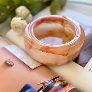 May include: A translucent, orange-toned resin bracelet. The bracelet has a rounded shape and a smooth surface. The color transitions from a light peach to a deeper orange hue. The bracelet is resting on a magazine.