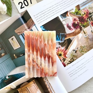 May include: A rectangular, decorative soap bar with a wavy, layered pattern in shades of red, orange, and cream. The soap is resting on an open magazine with text and images. A white tag with the number 207 is visible.