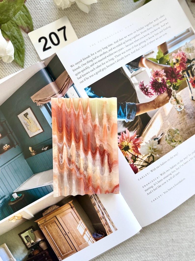 May include: A rectangular, decorative soap bar with a wavy, layered pattern in shades of red, orange, and cream. The soap is resting on an open magazine with text and images. A white tag with the number 207 is visible.