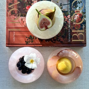 May include: Three circular, smooth stone bowls in shades of cream, pale pink, and marbled brown. The cream bowl holds a sliced fig, the pink bowl contains dark berries and a white flower, and the brown bowl holds a single yellow lemon.