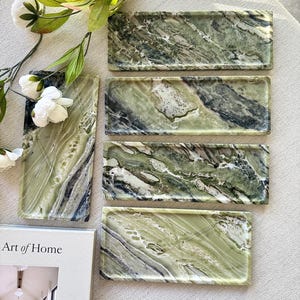 May include: Five rectangular trays crafted from green marble with distinct white and dark grey veining patterns. Each tray features a smooth, polished surface with slightly raised edges, showcasing natural stone variations in shades of olive and sage.