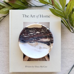 May include: A circular stone dish featuring a marbled pattern of deep brown, black, and white veins, resting on a light-colored hardcover book titled The Art of Home written by Shea McGee, surrounded by green leaves and white flower buds.