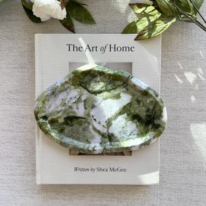 Puede incluir: Un plato decorativo de forma ovalada con un patrón veteado verde y blanco descansa sobre un libro titulado "The Art of Home" de Shea McGee. Flores blancas y hojas verdes en el fondo.