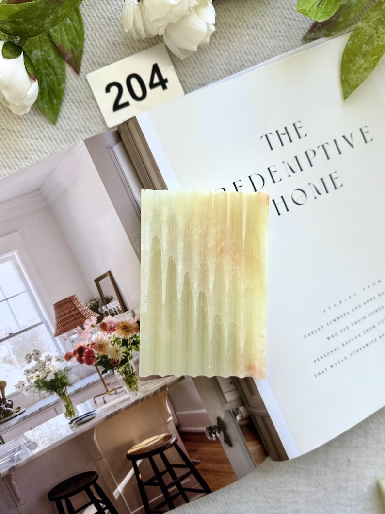 May include: A rectangular bar of soap with a wavy edge, featuring a light green and peach color gradient. The soap is placed on an open book with the title "THE REDEMPTIVE HOME" visible. A white tag with the number "204" is also present.