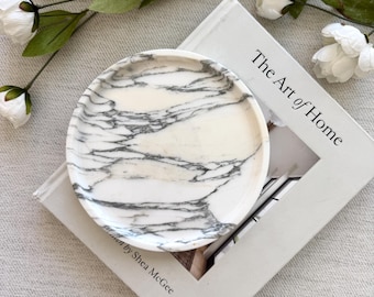 Pode incluir: Uma bandeja circular de mármore branco com veios cinzentos proeminentes, pousada sobre um livro intitulado The Art of Home de Shea McGee. A bandeja apresenta uma superfície lisa e polida com uma borda elevada, exibindo padrões de pedra.