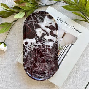 May include: An oval-shaped tray featuring a deep burgundy and white marbled pattern with a glossy finish. The tray is positioned on top of a white book titled The Art of Home, accompanied by a decorative branch with green leaves and white flowers.