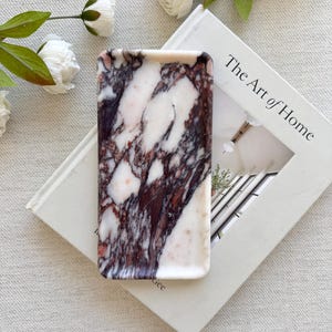 May include: A rectangular marble tray with a white and burgundy pattern. The tray is resting on a book titled "The Art of Home" with white flowers in the background. The tray is a decorative item.