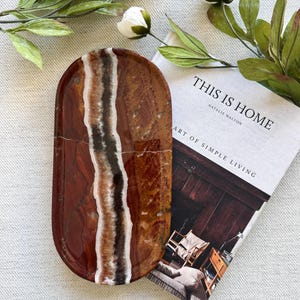 Pode incluir: Uma bandeja de formato oval feita de pedra polida em um tom marrom-avermelhado profundo com uma faixa vertical proeminente e texturizada de camadas minerais brancas, bronzeadas e marrom-escuras que atravessa o centro da superfície lisa.