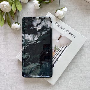May include: A rectangular phone case with a dark green and white marble design. The case is resting on a white book titled "The Art of Home." White flowers and green leaves are arranged around the case and book.