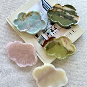 Peut inclure: Cinq plateaux décoratifs en forme de nuage de différentes couleurs et motifs. Les plateaux sont en pierre et reposent sur un livre. Les couleurs incluent le bleu clair, le vert, le rose et la crème. Le livre porte le mot "IFUL".