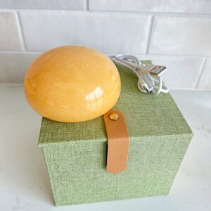 May include: An orange-yellow, spherical object sits atop a green box with a leather strap closure. A white USB charging cable is next to the sphere. The box has a textured fabric appearance.