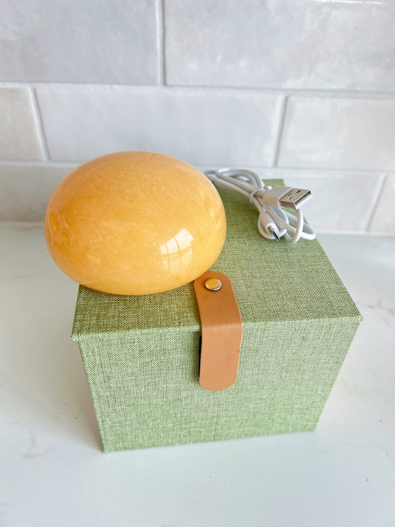 May include: An orange-yellow, spherical object sits atop a green box with a leather strap closure. A white USB charging cable is next to the sphere. The box has a textured fabric appearance.