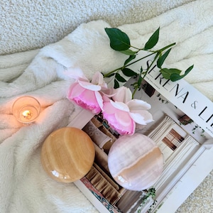 May include: Two polished, round stones in shades of beige and pink rest on a magazine with the text "SERENA & LILY". Pink roses and a lit candle are also present, all set against a soft, white, textured background.