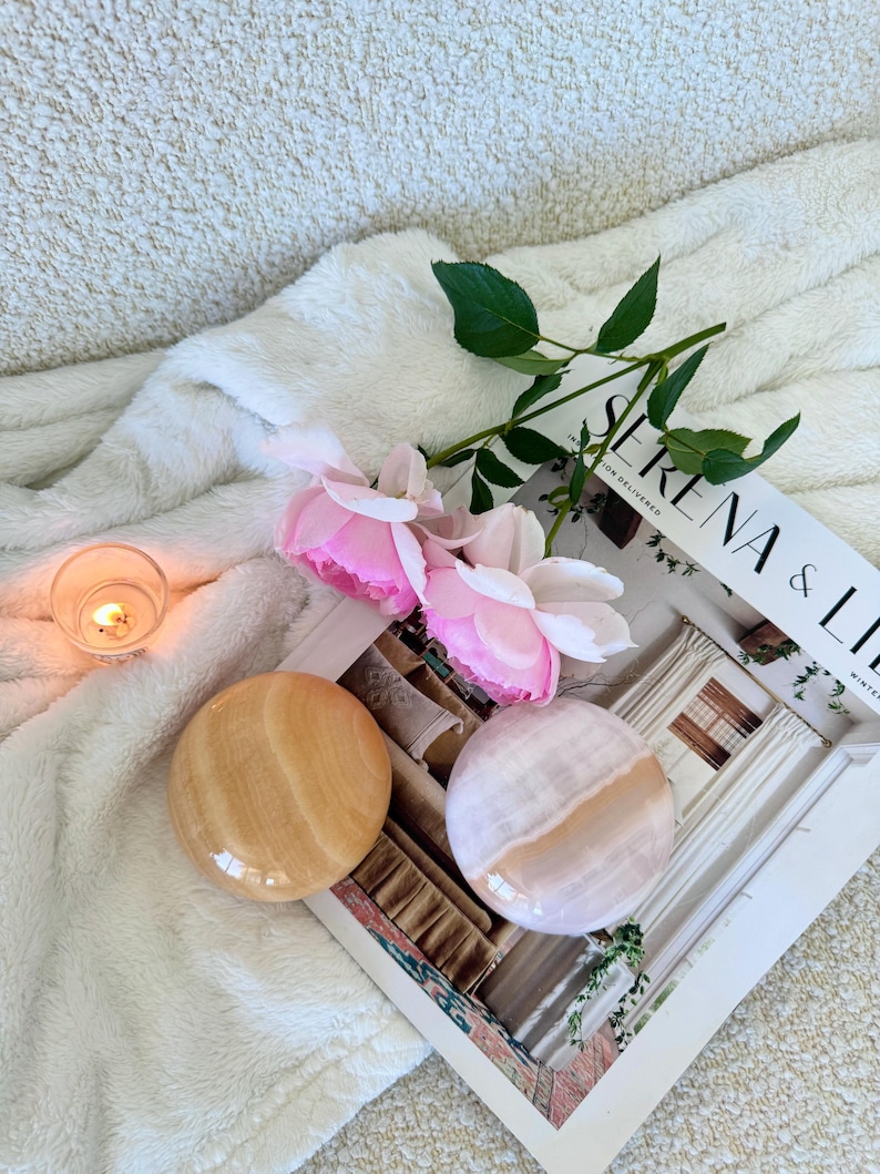 May include: Two polished, round stones in shades of beige and pink rest on a magazine with the text "SERENA & LILY". Pink roses and a lit candle are also present, all set against a soft, white, textured background.