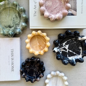 Puede incluir: Una colección de seis bandejas circulares de piedra con bordes festoneados en forma de burbuja. Las bandejas están hechas de varias piedras naturales, incluyendo mármol verde, rosa, amarillo, negro y blanco, dispuestas alrededor de dos libros.