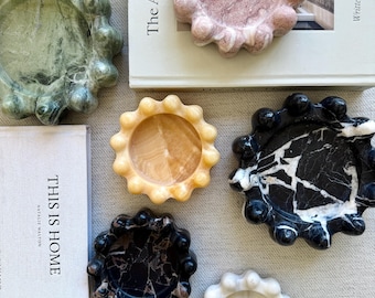 Puede incluir: Una colección de seis bandejas circulares de piedra con bordes festoneados en forma de burbuja. Las bandejas están hechas de varias piedras naturales, incluyendo mármol verde, rosa, amarillo, negro y blanco, dispuestas alrededor de dos libros.
