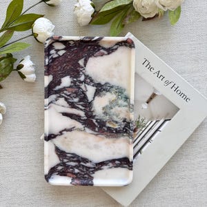 May include: A rectangular marble tray with a white, burgundy, and green pattern. The tray is next to a book titled "The Art of Home" and white flowers with green leaves.