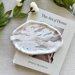 May include: An oval-shaped tray with a marbled white and gray pattern resting on a book titled The Art of Home written by Shea McGee. The tray has a smooth, polished surface with organic, flowing veins of gray color throughout the white base.