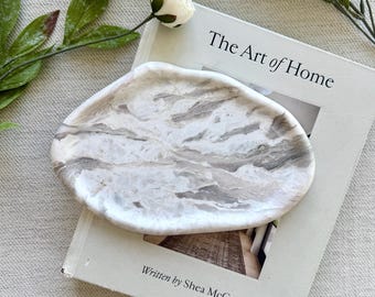 May include: An oval-shaped tray with a marbled white and gray pattern resting on a book titled The Art of Home written by Shea McGee. The tray has a smooth, polished surface with organic, flowing veins of gray color throughout the white base.