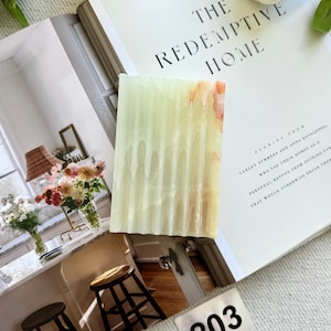 May include: A rectangular, light green and orange-toned soap bar with vertical ridges. The soap is displayed on an open book titled "THE REDEMPTIVE HOME". A small metal tag with the number "203" is visible.